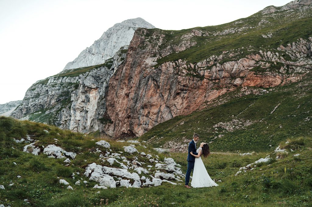 Sesja ślubna w Słowenii Młoda Pani w białej sukni ślubnej obejmująca Młodego Pana w granatowym garniturze na tle gór na Słowenii."