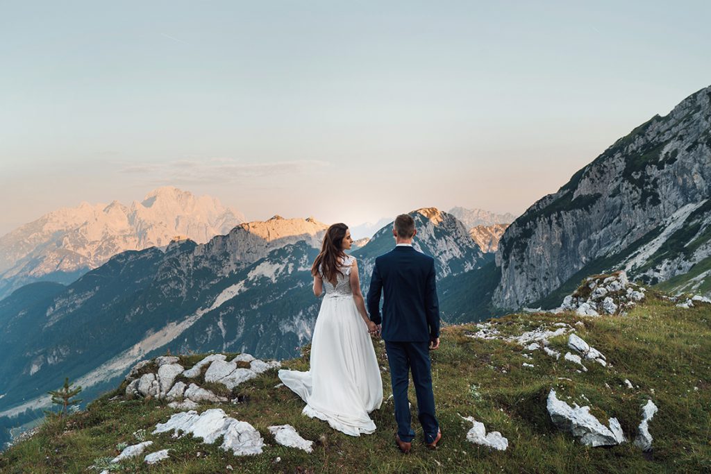 "Młoda Pani w sukni ślubnej i Młody Pan w garniturze stojący tyłem na tle gór na Słowenii."