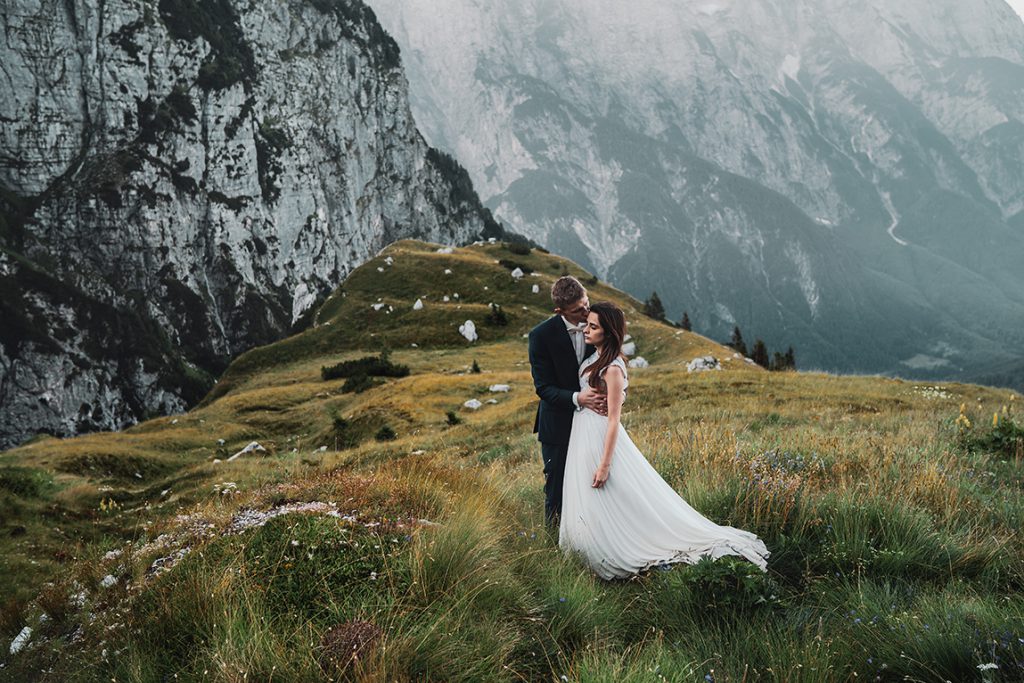 Sesja ślubna w Słowenii Młody Pan całujący Młodą Panią w czoło na tle mglistych gór w Alpach Julijskich."