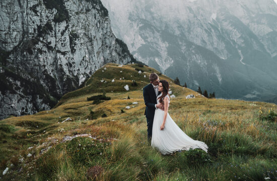 Sesja ślubna w Słowenii Młody Pan całujący Młodą Panią w czoło na tle mglistych gór w Alpach Julijskich.